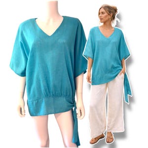 May include: A turquoise V-neck top with short sleeves and a tie detail at the waist. The top is made of a lightweight fabric and is paired with cream-colored wide-leg pants. The outfit is displayed on a mannequin and a person.