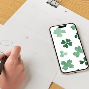 May include: A person writing on a white calendar with a black pen. A smartphone with a green shamrock pattern rests on the calendar. A black binder clip and a green notepad are also visible.