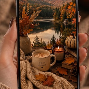 Op de afbeelding: Een smartphone die een digitale afbeelding van een herfstlandschap met een meer, bergen en bomen weergeeft. Het scherm toont een beige mok met het opschrift COZY SEASON, een kaars met het opschrift PUMPKIN SPICE, een pompoen en herfstbladeren.