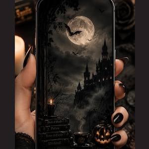 May include: A smartphone displaying a digital wallpaper of a gothic castle under a full moon with flying bats. The foreground features a stack of books labeled Book of Spells, Potions, Hexes & Curses, and Witchcraft, a steaming cup, and a glowing jack-o'-lantern.