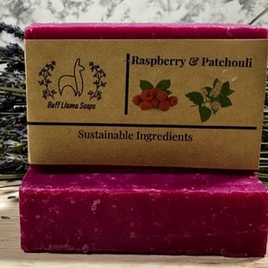 May include: A bar of deep pink soap with a brown label that reads "Raspberry & Patchouli" and "Buff Llama Soaps". The label also features a small illustration of a llama and the text "Sustainable Ingredients".