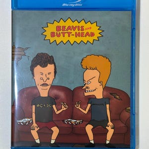 May include: A blue Blu-ray disc case featuring the animated characters Beavis and Butt-Head. The cover art shows the two characters sitting on a red couch. The text "BEAVIS AND BUTT-HEAD" is in a yellow starburst. Beavis wears an AC/DC shirt.