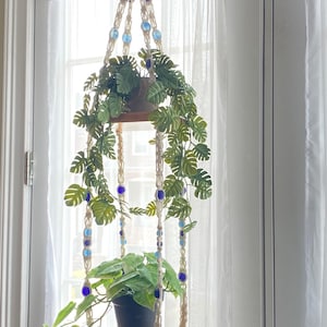 May include: A macrame plant hanger with two wooden shelves, adorned with blue and clear beads. The hanger supports two potted plants with green foliage. The design is suspended in front of a window with white curtains.