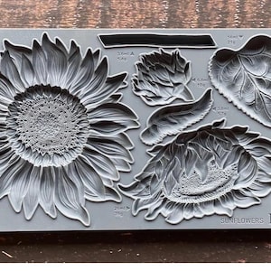 May include: A gray silicone mold with the words "SUNFLOWERS IOD" at the bottom. The mold has five different designs: a large sunflower, a smaller sunflower, three leaves, and a small sunflower bud. Each design has measurements in milliliters and grams.