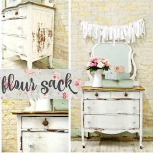 May include: A white antique dresser with a distressed finish and a wood top. The dresser has four drawers and is decorated with a white fabric garland with fringe. The dresser is sitting in front of a brick wall.