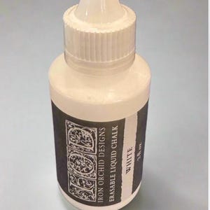 May include: A white plastic squeeze bottle of Iron Orchid Designs erasable liquid chalk. The black label features the text IOD, Iron Orchid Designs, Erasable Liquid Chalk, White, and 2 fl oz. The bottle has a white screw-on cap.