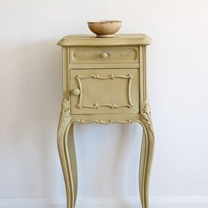 May include: A small, ornate, light brown side table with a single drawer and a door. The table has carved details and sits on four legs. A small, brown bowl sits on top of the table.