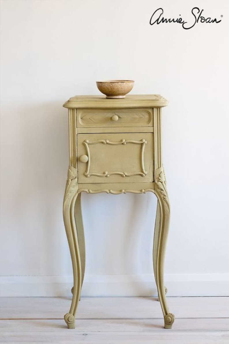 May include: A small, ornate, light brown side table with a single drawer and a door. The table has carved details and sits on four legs. A small, brown bowl sits on top of the table.