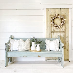 May include: A light blue wooden bench with two white pillows and two white ceramic pitchers with lavender flowers. The bench is in front of a white shiplap wall and a wooden door with a cotton wreath.