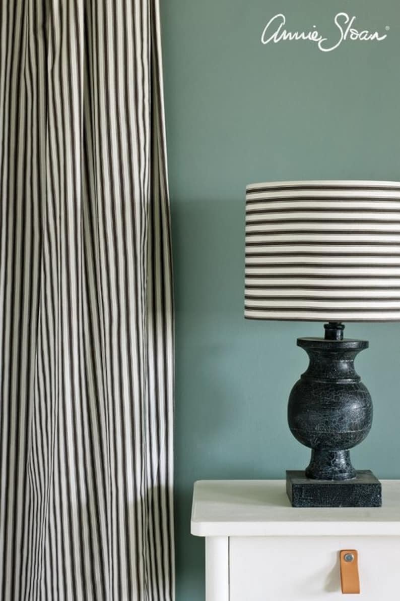 May include: A black and white striped lampshade sits atop a black lamp base with a distressed finish. The lamp is on a white nightstand with a brown leather drawer pull. The wall behind the lamp is painted a light teal green.