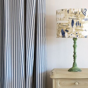 May include: A white nightstand with a green lamp with a patterned lampshade. The lampshade has a beige background with blue and white abstract designs. The nightstand has a drawer and a door with a decorative handle. The wall behind the nightstand is white and has a blue and white striped curtain.