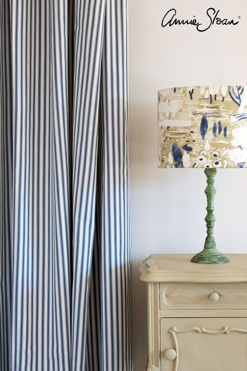 May include: A white nightstand with a green lamp with a patterned lampshade. The lampshade has a beige background with blue and white abstract designs. The nightstand has a drawer and a door with a decorative handle. The wall behind the nightstand is white and has a blue and white striped curtain.
