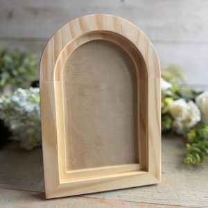 May include: A light-colored, unfinished wooden arch-shaped frame. The frame has a recessed area within the arch, and the wood grain is visible. The frame is likely for displaying photos or artwork. The background includes white flowers and greenery.