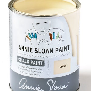 May include: A can of Annie Sloan Chalk Paint in the color Cream. The can is white with a gray label featuring a handprint design and the text "Annie Sloan Paint" and "Chalk Paint".
