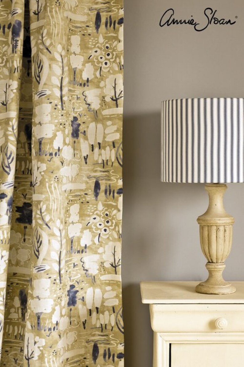 May include: A close-up of a fabric curtain with a white and blue abstract pattern. The curtain is hanging in front of a light gray wall. A lamp with a striped lampshade is on a white nightstand. The lampshade is white with blue stripes. The nightstand is white with a drawer. The text "Annie Sloan" is written in black on the wall.
