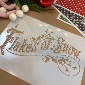 May include: A rectangular craft stencil featuring the text Flakes of Snow in a decorative script font. The design includes ornate flourishes and leaf-like accents integrated into the lettering. The stencil is made of a thin, semi-transparent material.