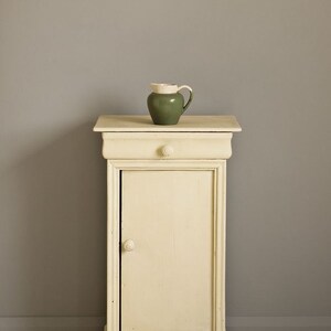 May include: A small, white painted cabinet with a single drawer and door. The cabinet is sitting on a light wood floor in front of a gray wall. A small green pitcher sits on top of the cabinet.