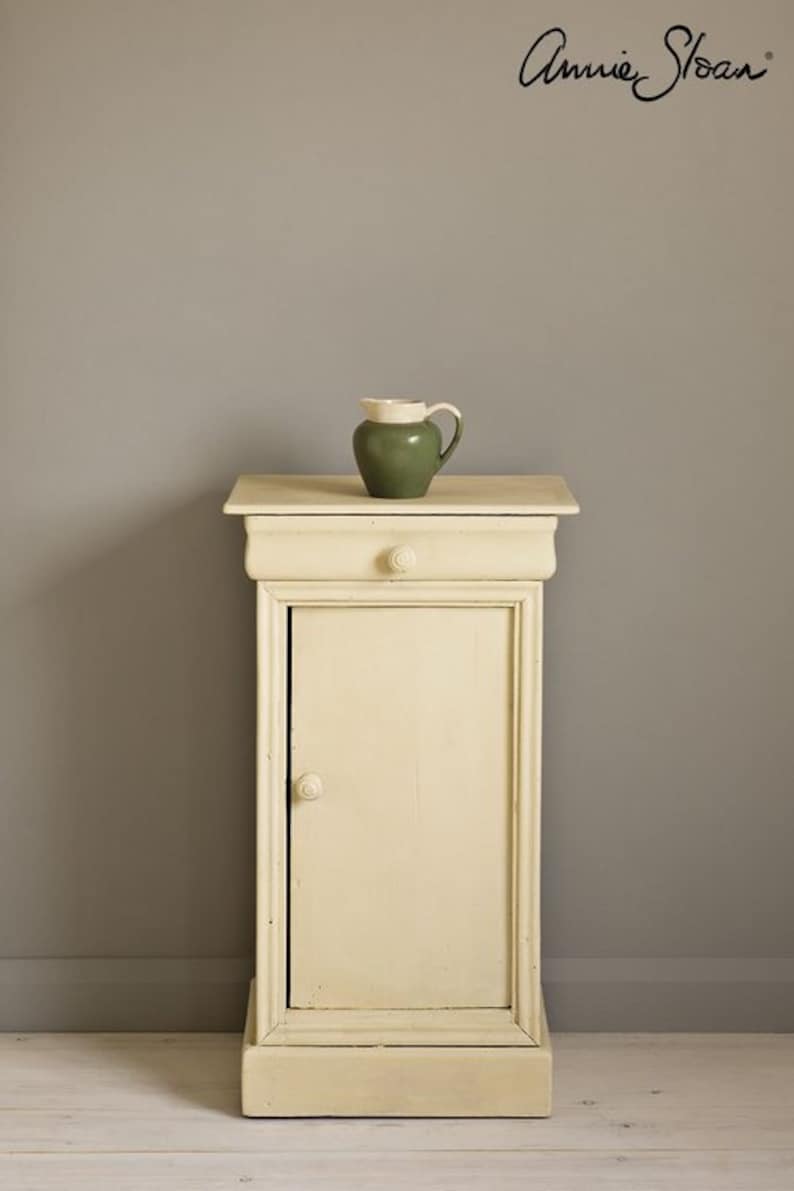 May include: A small, white painted cabinet with a single drawer and door. The cabinet is sitting on a light wood floor in front of a gray wall. A small green pitcher sits on top of the cabinet.