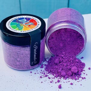 May include: Two clear jars of purple powder, one upright and one tipped over, spilling the vibrant pigment. The jars have black lids and labels with text. The spilled powder creates a small pile on a white surface.