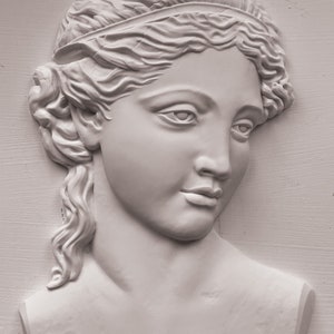 May include: A white plaster bust of a woman with a headband and long hair. The bust is sculpted in a classical style and has a serene expression.