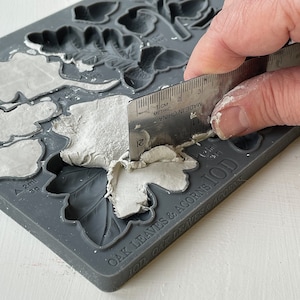 May include: A gray silicone mold with the text "OAK LEAVES & ACORNS IOD OAK LEAVES & ACORNS" printed on the bottom. The mold has several leaf and acorn shapes. A person is using a metal ruler to smooth out white clay in the mold.