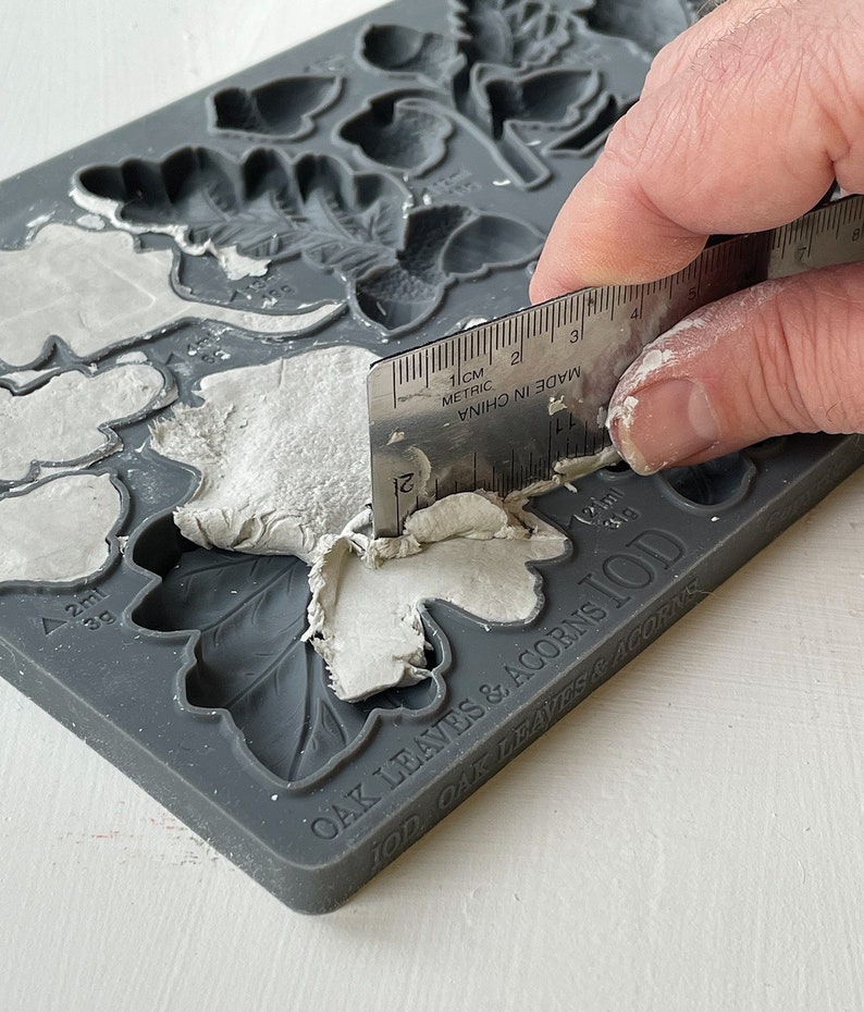 May include: A gray silicone mold with the text "OAK LEAVES & ACORNS IOD OAK LEAVES & ACORNS" printed on the bottom. The mold has several leaf and acorn shapes. A person is using a metal ruler to smooth out white clay in the mold.