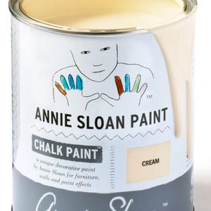 May include: A can of Annie Sloan Chalk Paint in the color Cream. The can is gray with a white label and features a handprint design. The label reads "Annie Sloan Paint" and "Chalk Paint".