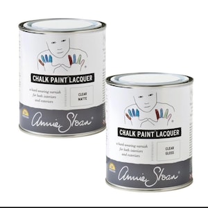 May include: Two cylindrical metal cans of Annie Sloan Chalk Paint Lacquer. One can is labeled Clear Matte and the other is labeled Clear Gloss. Both white cans feature a line drawing of a person with colorful painted fingers and UV protection.