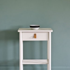 May include: White side table with a single drawer and a lower shelf. The drawer has a leather pull. The table is against a teal wall. Annie Sloan is written in cursive on the wall.