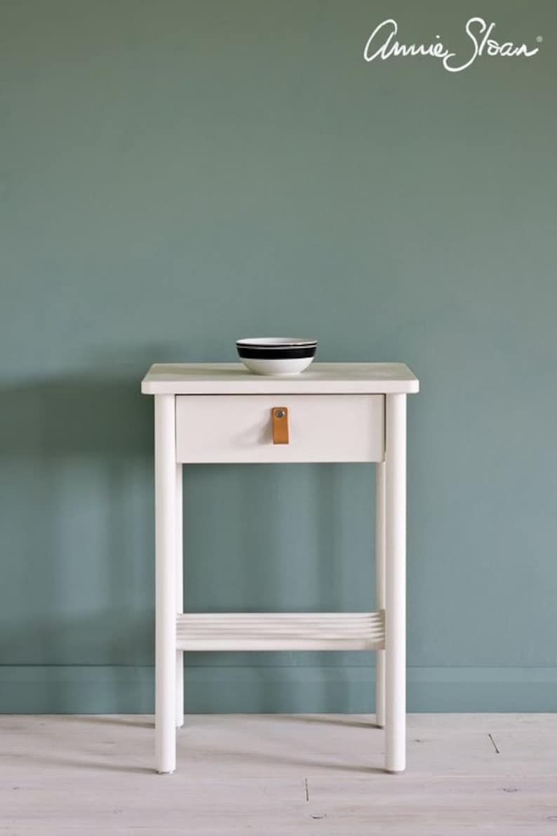 May include: White side table with a single drawer and a lower shelf. The drawer has a leather pull. The table is against a teal wall. Annie Sloan is written in cursive on the wall.