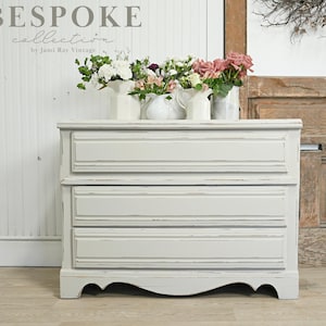 May include: A white three-drawer dresser with a distressed finish. The dresser has a curved base and is made of wood. The dresser is sitting on a wooden floor. The dresser is decorated with flowers in white and pink vases.