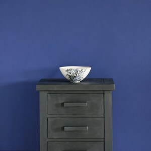 May include: A dark gray wooden nightstand with three drawers and black drawer pulls. The nightstand is sitting on a white wooden floor in front of a wall painted a deep blue color. The text "Annie Sloan" is written in white on the wall.