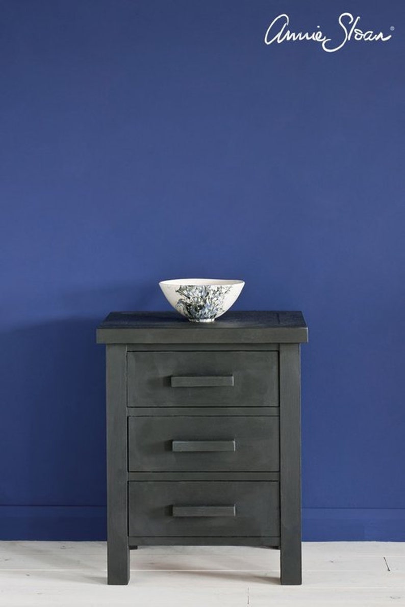 May include: A dark gray wooden nightstand with three drawers and black drawer pulls. The nightstand is sitting on a white wooden floor in front of a wall painted a deep blue color. The text "Annie Sloan" is written in white on the wall.