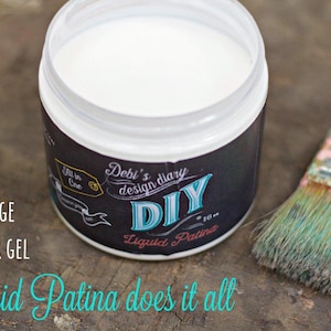 May include: A white jar of Debi's design diary DIY Liquid Patina. The jar has a black label with white text. The text on the label reads "Debi's design diary DIY Liquid Patina". The jar is sitting on a wooden surface next to a paintbrush.