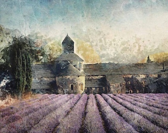 May include: A digital art print depicting a stone monastery with a central tower and dark slate roof, situated behind rows of vibrant purple lavender fields. The scene features textured brushstrokes, muted green trees, and a soft, hazy sky overhead.