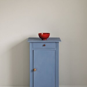May include: A small blue painted cabinet with a single drawer and a door. The cabinet has a wooden knob on the door and a small red bowl on top.