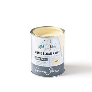 May include: A white can of Annie Sloan Chalk Paint in the color Cream. The can has a label with a cartoon illustration of a person with colorful fingertips. The label also includes the text "Annie Sloan Paint" and "Chalk Paint".