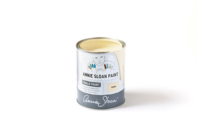 May include: A white can of Annie Sloan Chalk Paint in the color Cream. The can has a label with a cartoon illustration of a person with colorful fingertips. The label also includes the text "Annie Sloan Paint" and "Chalk Paint".