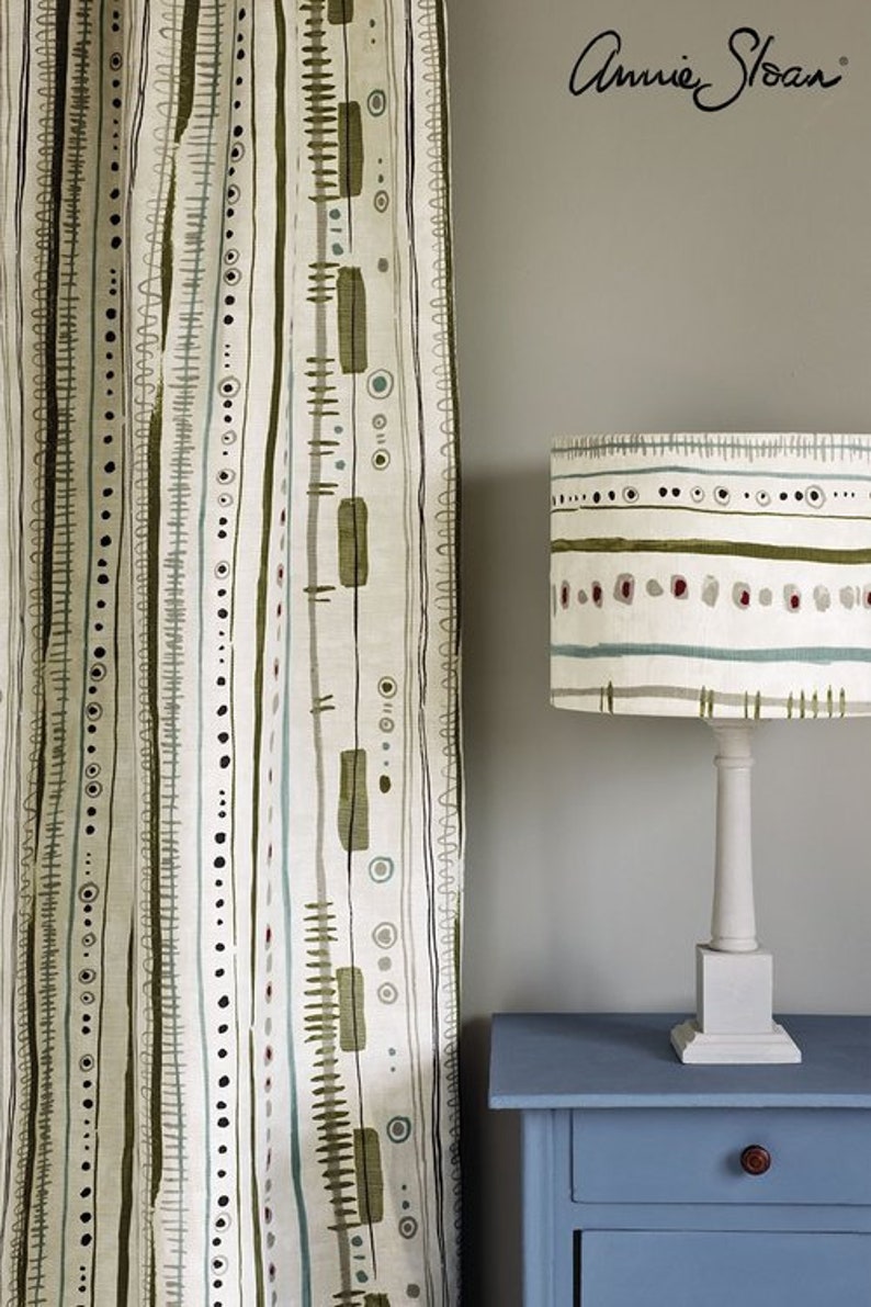 May include: A white lampshade with a green, brown, and blue geometric pattern. The lampshade is on a white lamp base and sits on a blue table with a drawer. The text "Annie Sloan" is visible in the upper right corner of the image.