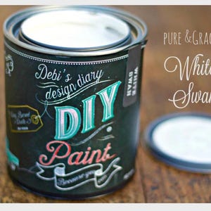 May include: A white paint can with a black label that says "Debi's design diary DIY Paint". The label also has a green and pink design. The can is sitting on a wooden surface. The text "PURE & GRACEFUL White Swan" is visible in the background.