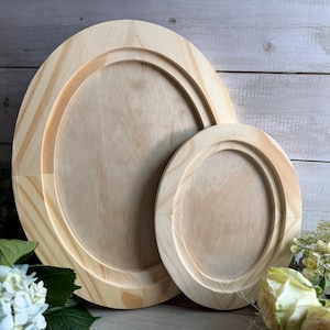 May include: Two unfinished wooden oval frames, one large and one small, are displayed. The frames have a natural wood color and a raised, rounded edge. The background features a light-colored wooden surface and greenery.