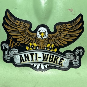 Op de afbeelding: Een sticker met een zwarte en gouden adelaar met gespreide vleugels. De adelaar houdt een banner vast waarop "ANTI-WOKE" in gele letters staat. De sticker heeft een klein logo in de linkerbenedenhoek.