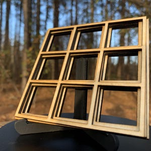 May include: A wooden decorative window frame with nine square openings. The frame is made of light-colored wood and is displayed on a black stand against a blurred outdoor background. The frame is approximately 6 inches wide and 6 inches tall.