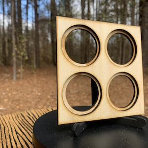 May include: A square, unfinished wooden organizer with four circular cutouts. The organizer is displayed on a black stand, with a blurred forest background. The wood has a light, natural color.