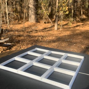 May include: A white, rectangular grid with twelve evenly spaced square compartments. The grid is made of a solid material and sits on a dark surface. The background features a natural outdoor setting with trees and foliage.