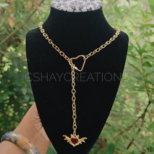 May include: A gold-toned lariat necklace featuring a heart-shaped carabiner clasp and a dangling charm shaped like a winged heart with a red and black checkered pattern. The text CSHAYCREATIONS is visible across the center of the display.
