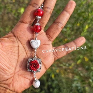 May include: A long, dangling earring featuring red beads, a white heart-shaped stone, and a red rose pendant. The earring is silver-toned and has a white, pear-shaped bead at the bottom. The text "CSHAYCREATIONS" is visible.