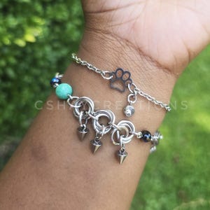 May include: Two silver-toned chain bracelets. The top bracelet features a small paw print charm and a clear crystal. The bottom bracelet consists of interlocking metal rings, three small silver spikes, and turquoise and dark blue beads.