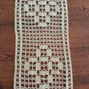 May include: A beige crocheted table runner with a diamond pattern. The runner is on a wooden surface.