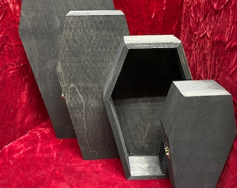 May include: A set of four wooden boxes shaped like coffins with a dark charcoal finish. Three of the boxes are closed, while one is open to reveal a hollow interior. Each box features a small gold-colored metal latch on the side.